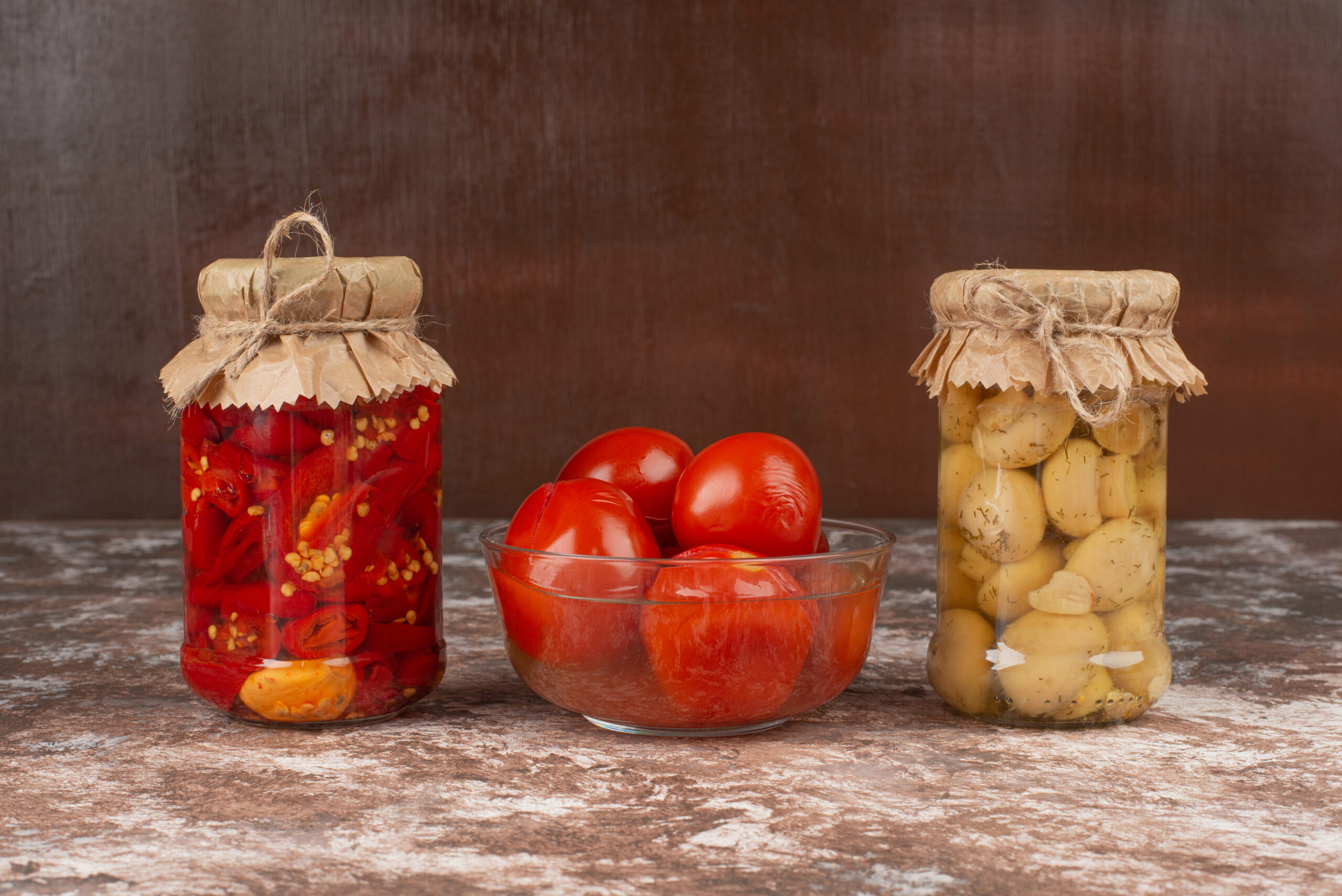 Pickled red peppers and mushrooms in a glass jar on marble table with bowl of pickled tomatoes. High quality photo